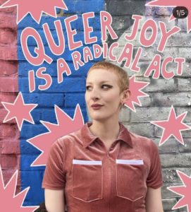 Bee poses in front of a brick wall surrounded by text that reads Queer joy is a radical act. 