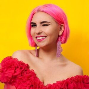 Ruby smiles and winks at the camera, she wears a red off the shoulder top, has pink hair, and is in front of a yellow background. Diversify